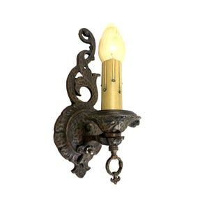 May include: An antique wall sconce with a decorative, ornate design. The sconce features a bronze finish, a candle-shaped light bulb, and a detailed, scrollwork backplate. The light bulb is lit, casting a warm glow.