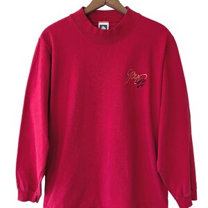 May include: A long-sleeved, pink mock neck t-shirt with a small embroidered logo on the front. The logo is a stylized "Joy" with a small graphic of a bird.