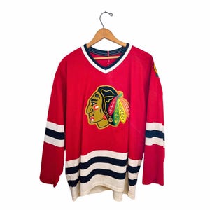 May include: Red hockey jersey with the Chicago Blackhawks logo on the front. The jersey has white and black stripes on the sleeves and bottom hem. The V-neck collar is white with a black trim. The logo features a Native American head.