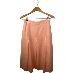 May include: A peach-colored skirt with pleats, hanging on a wooden hanger. The skirt has a textured appearance and a classic design. The garment is made of a lightweight fabric, suitable for warmer weather.