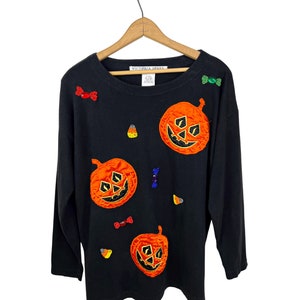 May include: Black long-sleeved shirt with three orange pumpkin appliques and colorful candy embellishments. The shirt has the text "Victoria Jane" on the tag.