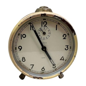 May include: Vintage cream-colored alarm clock with a gold-tone bezel and black hands. The clock face has black numerals and the word "ALARM" is visible. The clock is marked "GERMANY" at the bottom.
