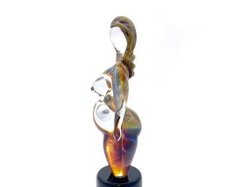 May include: A glass sculpture of a pregnant woman with a clear body and a brown, orange, and purple iridescent base. The sculpture is standing on a black pedestal.
