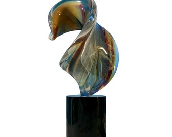 Könnte beinhalten: Eine farbenfrohe Glasskulptur mit einem wirbelnden, abstrakten Design. Die Skulptur zeigt Farbtöne von Blau, Braun und Gold, montiert auf einem schwarzen zylindrischen Sockel. Ein dekoratives Kunstwerk.