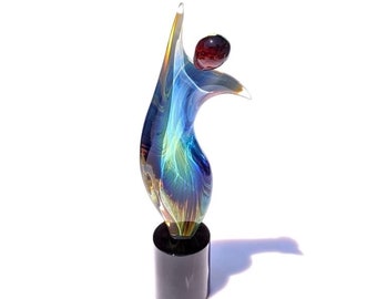Könnte beinhalten: Eine blaue und grüne Glasskulptur mit einer roten Glaskugel oben. Die Skulptur steht auf einem schwarzen Sockel.