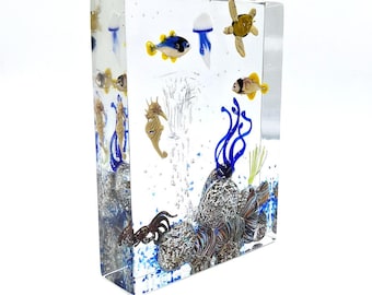 May include: A glass paperweight with a blue, white, and yellow jellyfish, a brown sea turtle, a blue and yellow fish, a brown seahorse, and blue coral in a clear glass block.