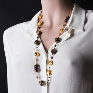 Può includere: Una lunga collana di perline di vetro dorate e marroni di diverse forme e dimensioni. La collana è su una camicia bianca.