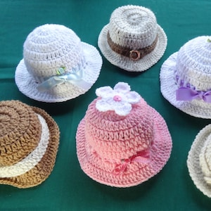 May include: A collection of seven handmade crochet hats in various colors, including white, brown, and pink. Some hats feature decorative ribbons and flowers. The hats are arranged on a green surface, showcasing their intricate designs.