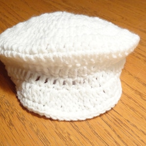 Könnte beinhalten: Eine weiße gehäkelte Mütze mit einem Rand. Die Mütze ist mit einem einfachen Stichmuster gearbeitet und hat eine abgerundete Krone.
