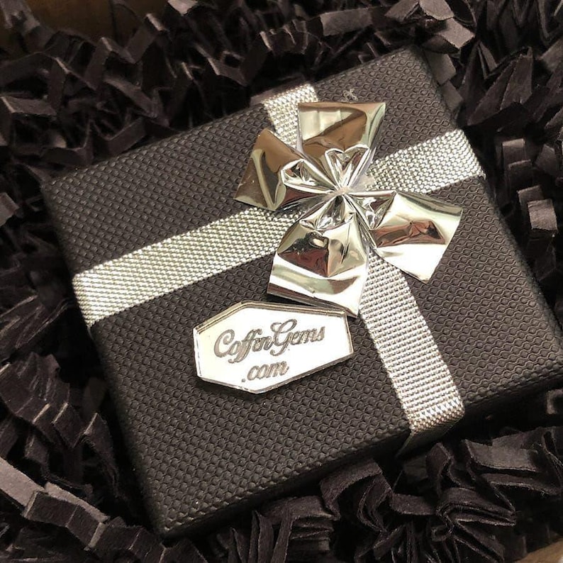 May include: A black gift box with a silver ribbon and bow. The box has a silver label with the text "Coffin Gems .com" on it.