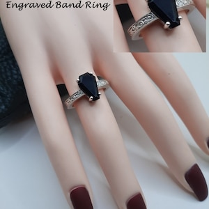 Könnte beinhalten: Ein silberner Bandring mit einem gravierten Design und einem schwarzen, sargförmigen Edelstein. Der Ring befindet sich an einem Modellfinger.