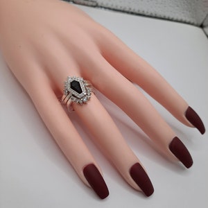 May include: A silver ring with a black gemstone surrounded by smaller clear gemstones. The ring is on a model's finger.