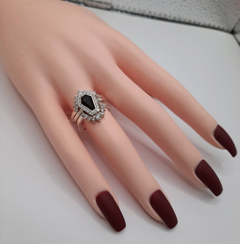 May include: A silver ring with a black gemstone surrounded by smaller clear gemstones. The ring is on a model's finger.