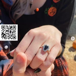 May include: A close-up of a hand wearing a silver ring with a black coffin-shaped stone. The ring is set with small diamonds around the stone.