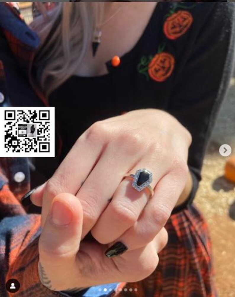 May include: A close-up of a hand wearing a silver ring with a black coffin-shaped stone. The ring is set with small diamonds around the stone.