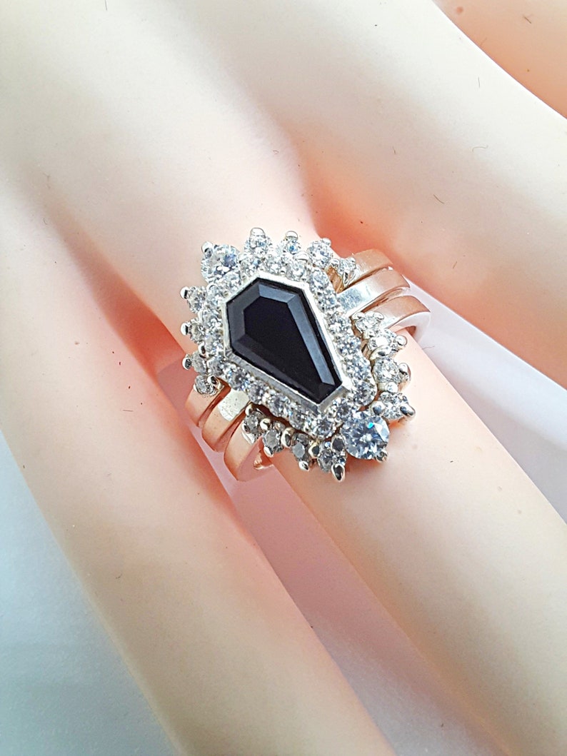 May include: A silver ring with a black coffin-shaped gemstone surrounded by clear cubic zirconia stones. The ring is set on a white background.