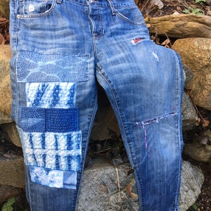 May include: Blue denim jeans with multiple patches sewn on the left leg. The patches are made of different fabrics and have a blue and white color scheme. The jeans have a distressed look with rips and tears.