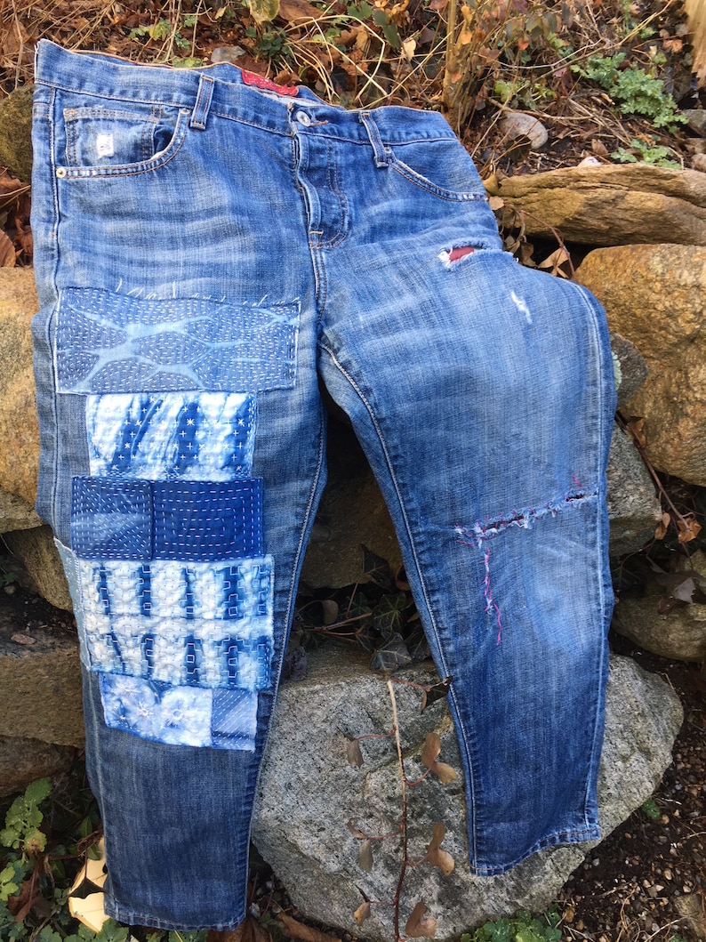 May include: Blue denim jeans with multiple patches sewn on the left leg. The patches are made of different fabrics and have a blue and white color scheme. The jeans have a distressed look with rips and tears.