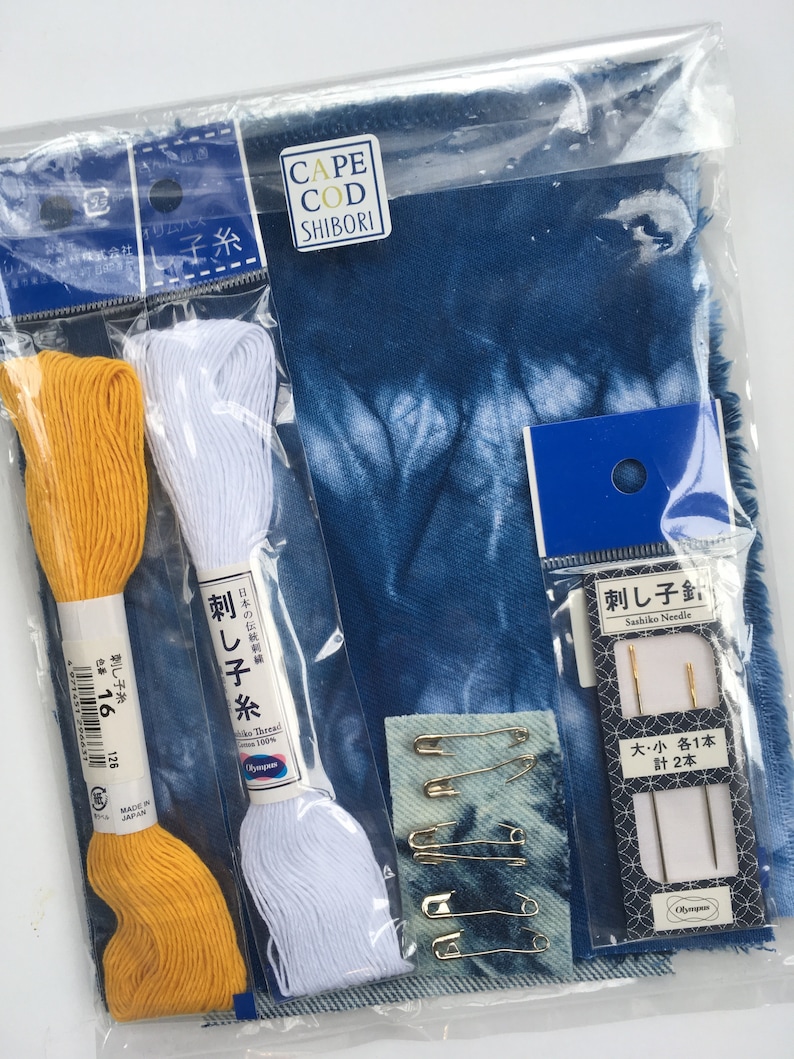 May include: A blue and white fabric kit for sashiko embroidery. The kit includes two spools of thread, one white and one yellow, three safety pins, two needles, and a piece of blue fabric with a shibori pattern. The packaging includes the text "Cape Cod Shibori" and "Sashiko Needle".