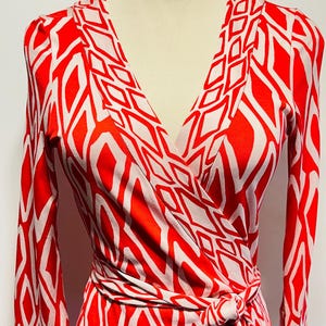 May include: A red and white patterned wrap dress with long sleeves. The dress features a V-neckline and a tie waist. The geometric pattern is a repeating design of white diamonds on a red background. The dress is displayed on a mannequin.