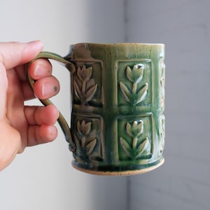 Peut inclure: Une tasse en céramique verte avec une anse et un motif floral en relief. La tasse est décorée d'un motif répétitif de tulipes.
