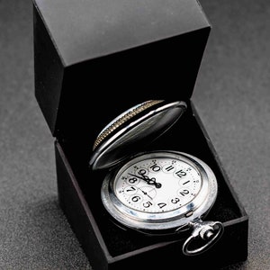 May include: A silver pocket watch with Roman numeral markings on the face, in a black velvet lined box.
