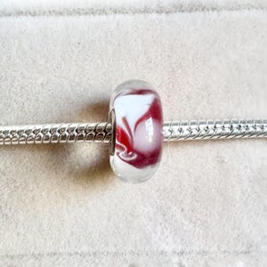 Può includere: Una perla cilindrica in vetro caratterizzata da un motivo a vortice nei colori bianco opaco e rosso intenso. La perla è infilata su un bracciale a catena snake color argento, con una superficie liscia e lucida e una finitura brillante.