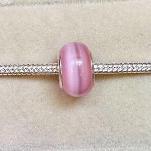 Può includere: Una perla rosa cilindrica e liscia con un effetto occhio di gatto cangiante, caratterizzata da un riflesso luminoso verticale centrale. La perla è infilata su un bracciale a catena snake color argento, dal design a maglie metalliche, flessibile e testurizzato.