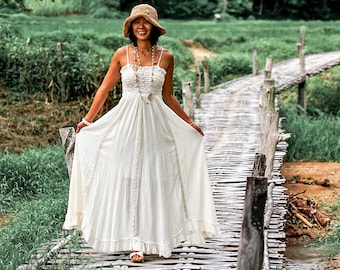 May include: A woman in a white maxi dress and a straw hat walks across a wooden bridge over a stream. The bridge is made of bamboo and is surrounded by lush green vegetation.