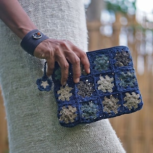 May include: A blue and beige crocheted bag with a square pattern. The bag is held by a person wearing a light gray knitted dress.