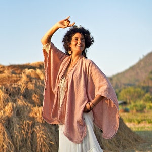 May include: A woman wearing a pink, loose-fitting,  boho-style top with a white, ruffled skirt. She is standing in a field of hay with a mountain in the background.