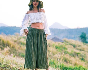 May include: A woman wearing a white off-the-shoulder crop top with ruffled sleeves and olive green striped wide-leg pants. She is also wearing a matching hat. The outfit is suitable for a casual, bohemian style.