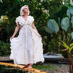 Puede incluir: Un vestido largo color crema con detalles de volantes. El vestido presenta un escote en V y mangas cortas con detalles de flecos. La modelo est&aacute; de pie descalza sobre una superficie de madera cerca de un cuerpo de agua, rodeada de exuberante vegetaci&oacute;n.