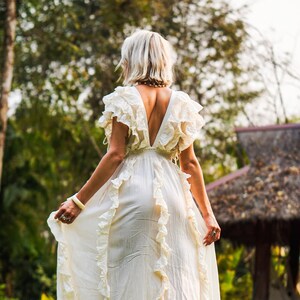 Puede incluir: Vestido largo color crema con volantes y escote en V pronunciado. El vestido est&aacute; hecho de una tela ligera y tiene una silueta fluida. La modelo est&aacute; de pie sobre escalones de piedra en un jard&iacute;n.