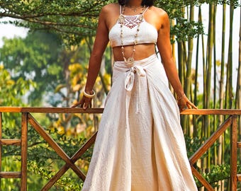 Puede incluir: Un conjunto de dos piezas color crema que consta de un top halter y pantalones de pernera ancha. El top presenta un diseño de ganchillo y los pantalones tienen un estilo envolvente. Un sombrero y sandalias a juego completan el conjunto. El atuendo se usa al aire libre.