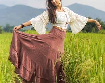 May include: A woman wearing a white peasant top with a brown tiered skirt and a beaded necklace stands in a field of tall green grass.