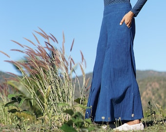 May include: A woman wearing a blue polka dot turtleneck shirt, a black hat, and blue wide-leg pants stands in a field of tall grass.