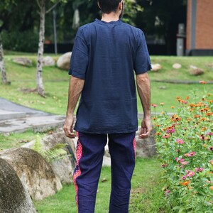 May include: A person wearing a dark blue short-sleeved shirt and dark blue pants with a pink and blue patterned trim. The person is walking away from the camera in a grassy area with flowers.