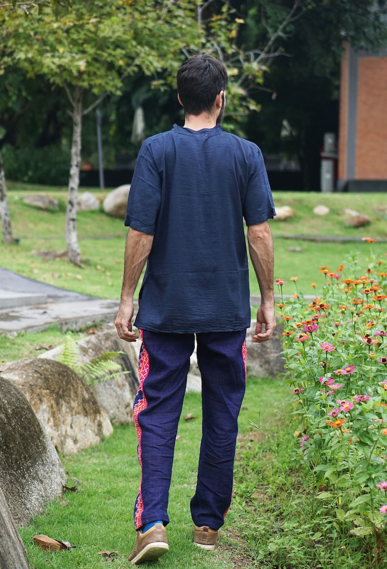 May include: A person wearing a dark blue short-sleeved shirt and dark blue pants with a pink and blue patterned trim. The person is walking away from the camera in a grassy area with flowers.