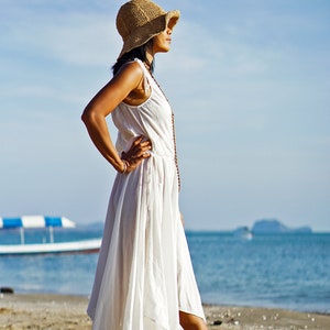 May include: A woman wearing a white maxi dress and a wide-brimmed straw hat stands on a sandy beach with the ocean in the background.