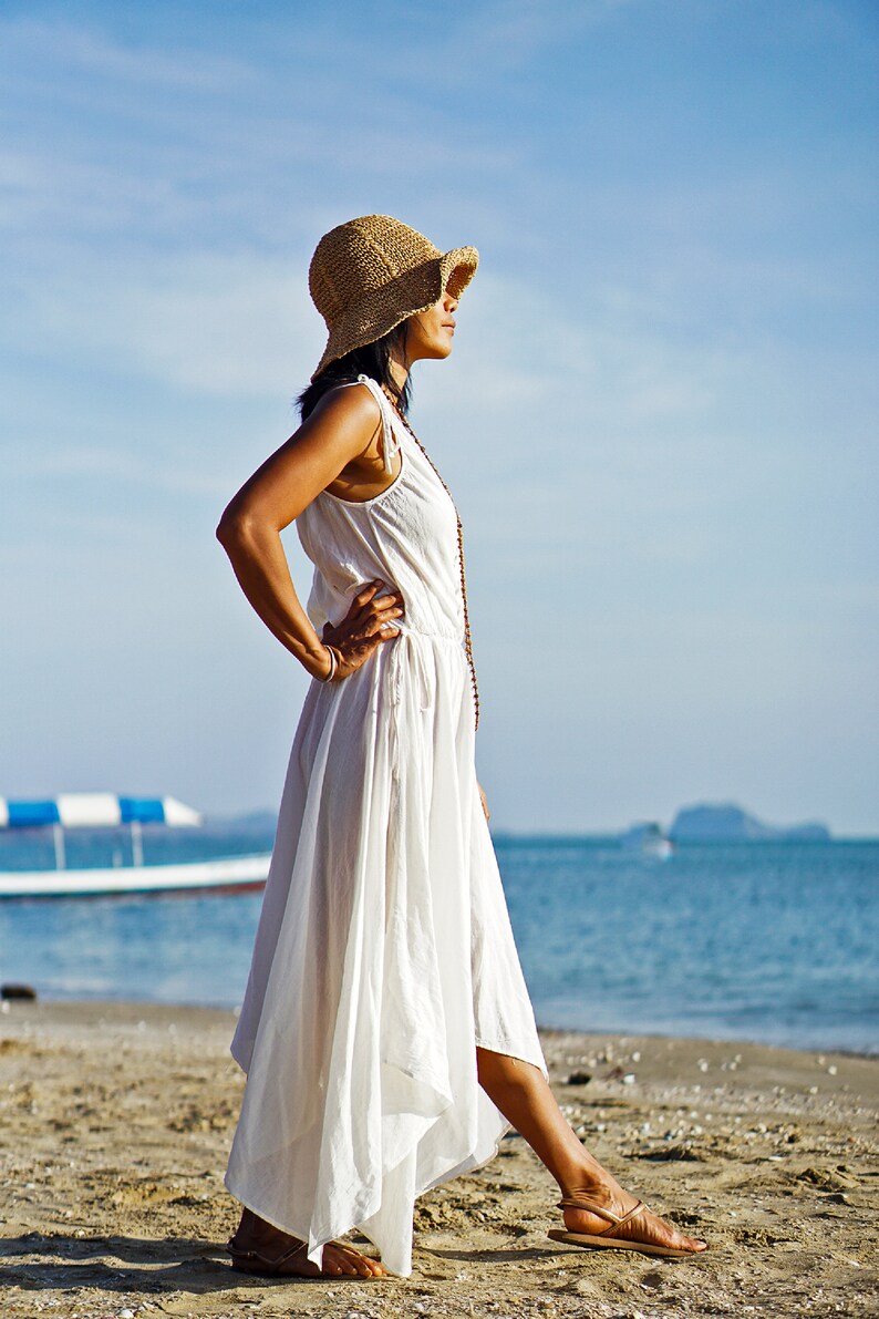 May include: A woman wearing a white maxi dress and a wide-brimmed straw hat stands on a sandy beach with the ocean in the background.