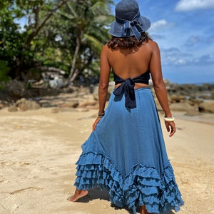 May include: A woman wearing a blue denim skirt with multiple ruffles, a black halter top tied at the back, and a blue sun hat. She is standing on a sandy beach.