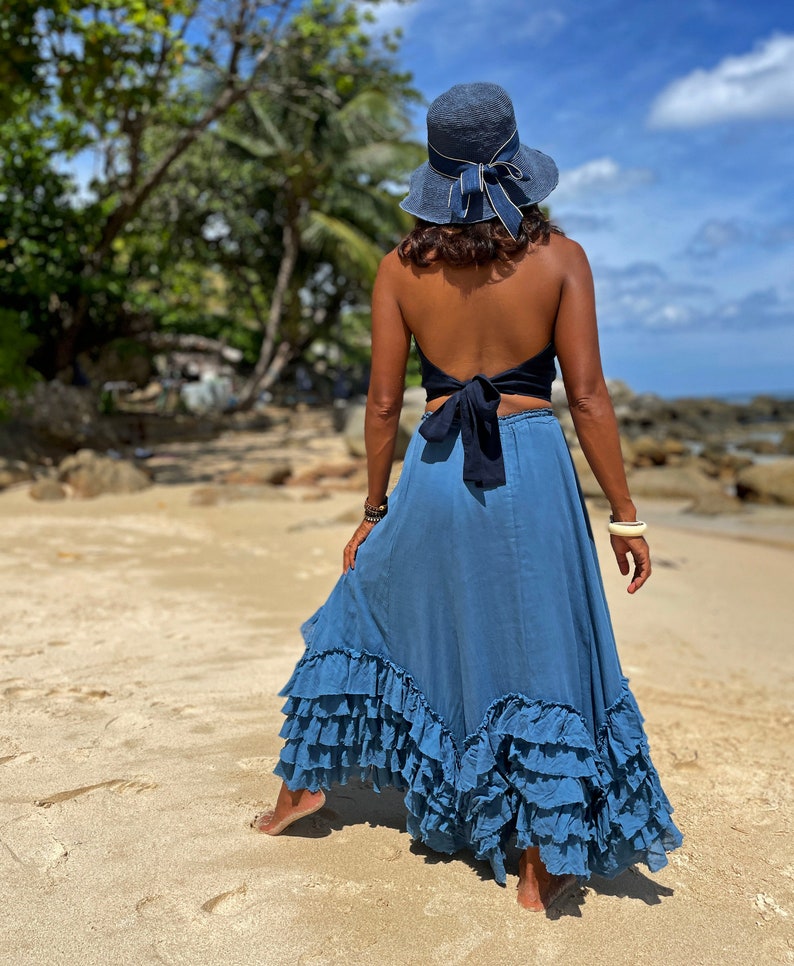 May include: A woman wearing a blue denim skirt with multiple ruffles, a black halter top tied at the back, and a blue sun hat. She is standing on a sandy beach.
