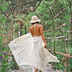 May include: A woman in a white halter dress and a sun hat walks along a wooden walkway under a metal archway covered in greenery. The dress has a flowing skirt with a patterned design.