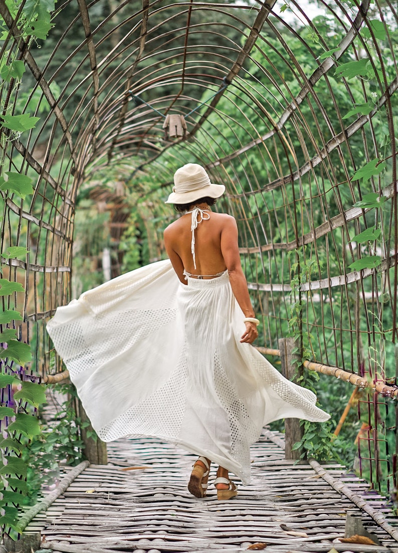 May include: A woman in a white halter dress and a sun hat walks along a wooden walkway under a metal archway covered in greenery. The dress has a flowing skirt with a patterned design.