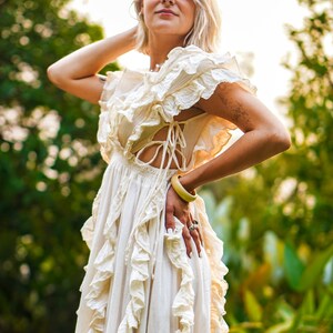 Puede incluir: Un vestido color crema con detalles de volantes y un corpi&ntilde;o con cordones. El vestido tiene una falda fluida y mangas cortas. Se ve un brazalete amarillo y anillos. El fondo es un follaje verde borroso.