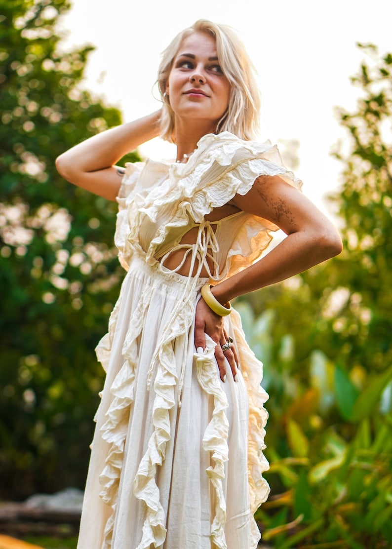 Puede incluir: Un vestido color crema con detalles de volantes y un corpi&ntilde;o con cordones. El vestido tiene una falda fluida y mangas cortas. Se ve un brazalete amarillo y anillos. El fondo es un follaje verde borroso.