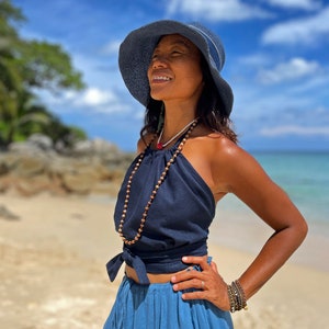 May include: A woman wearing a blue sun hat, a navy blue halter top with a brown beaded necklace, and a blue denim skirt. She is standing on a beach with a blue sky and white clouds in the background.