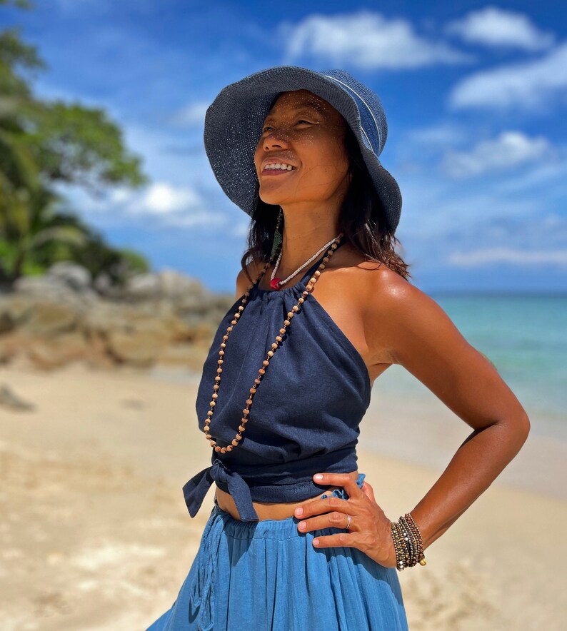 May include: A woman wearing a blue sun hat, a navy blue halter top with a brown beaded necklace, and a blue denim skirt. She is standing on a beach with a blue sky and white clouds in the background.
