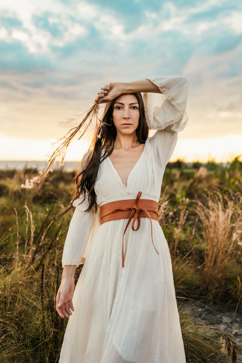 May include: A woman wearing a white dress with a brown leather belt stands in a field of tall grass. She is holding a long, thin branch in her hand and looking at the camera.